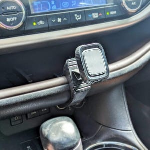 Può includere: Supporto nero per telefono da auto fissato al cruscotto. Il supporto ha una superficie magnetica rettangolare e un braccio flessibile. Il cruscotto dell'auto ha vari pulsanti e un display digitale.