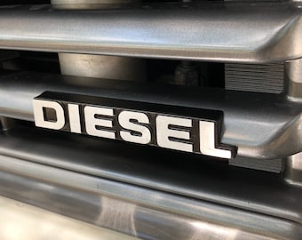 Może przedstawiać: Czarny i srebrny emblemat "DIESEL" na szarej metalowej kratce.