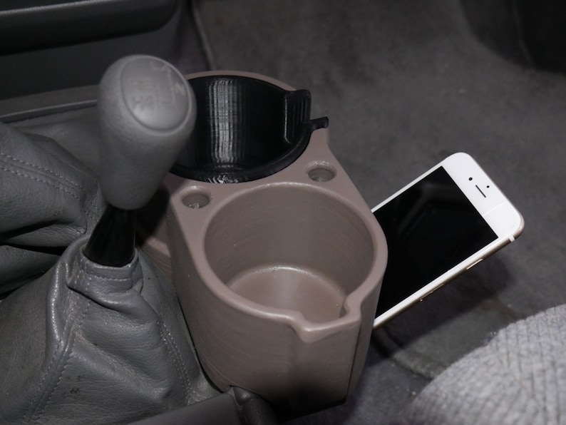 May include: A gray and black car cup holder with a phone holder attached to the side. The cup holder is made of plastic and has two cup holders. The phone holder is made of metal and has a spring-loaded clip to hold the phone in place.