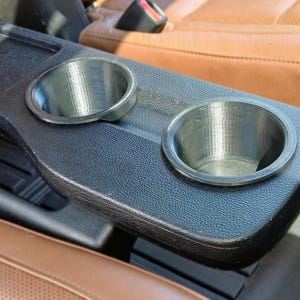 May include: A black plastic car console with two circular cup holders. The cup holders are silver and appear to be 3D-printed. The console is set against a background of brown leather car seats.