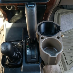 May include: A beige plastic cup holder with two cup slots, one black and one beige, is mounted to the centre console of a car. The cup holder is next to a black gear shift lever with a black knob and a gear shift indicator with the letters P, R, N, D, 2, and L.