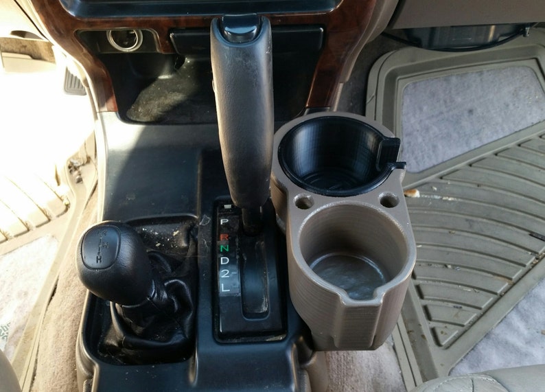 May include: A beige plastic cup holder with two cup slots, one black and one beige, is mounted to the centre console of a car. The cup holder is next to a black gear shift lever with a black knob and a gear shift indicator with the letters P, R, N, D, 2, and L.