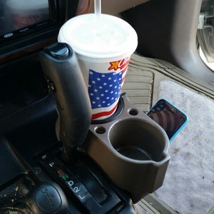 May include: A grey plastic cup holder with two compartments, one for a drink and one for a phone. A red, white and blue cup with a straw is in the drink holder. A blue phone with a red, white and blue screen is in the phone holder.