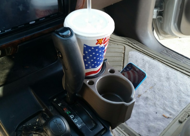 May include: A grey plastic cup holder with two compartments, one for a drink and one for a phone. A red, white and blue cup with a straw is in the drink holder. A blue phone with a red, white and blue screen is in the phone holder.