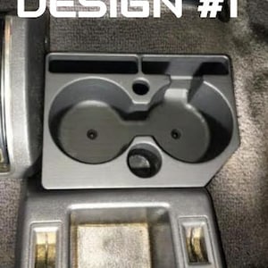 May include: A dark gray plastic automotive center console cup holder insert featuring two circular beverage compartments, a rectangular storage slot, and a small circular opening, with the text DESIGN #1 printed in white above the unit.
