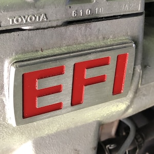 May include: Close-up of a silver engine part with a red "EFI" label. The label is rectangular and has a brushed metal finish. The engine part is made of metal and has a gray finish.