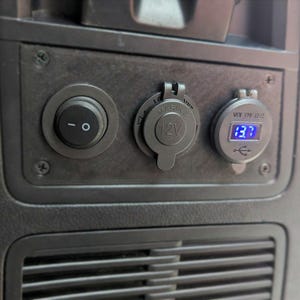 May include: Close-up of a black panel with a round power switch, a 12V outlet, and a USB port with a digital voltage display showing 13.7 volts. The panel is mounted in a vehicle interior.