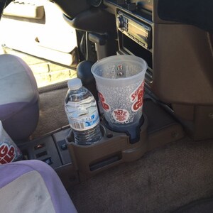 May include: A car's interior with a brown dashboard and a purple seat. There are two clear plastic cups with the text "Big Gulp" on them and a clear plastic bottle of water in the cup holders.