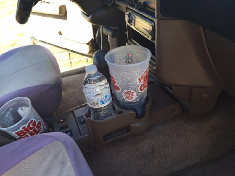 May include: A car's interior with a brown dashboard and a purple seat. There are two clear plastic cups with the text "Big Gulp" on them and a clear plastic bottle of water in the cup holders.