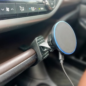 Könnte beinhalten: Ein schwarzer und grauer magnetischer Telefonhalter, der im Inneren eines Autos befestigt ist. Die Halterung hat einen kreisförmigen Kopf mit blau beleuchtetem Rand. Ein schwarzes Kabel erstreckt sich von der Halterung. Die Halterung ist an einer braunen Stange befestigt.