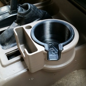 May include: A beige plastic cup holder with a black cup insert. The cup holder is mounted in a car's center console.