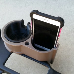 May include: A brown and black car cup holder with a phone holder on the side. A white phone with a black case is in the phone holder.