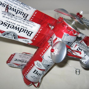 May include: A red, white, and blue Budweiser beer can airplane model with silver wings and propellers. The airplane has red and silver accents and is decorated with rhinestones.