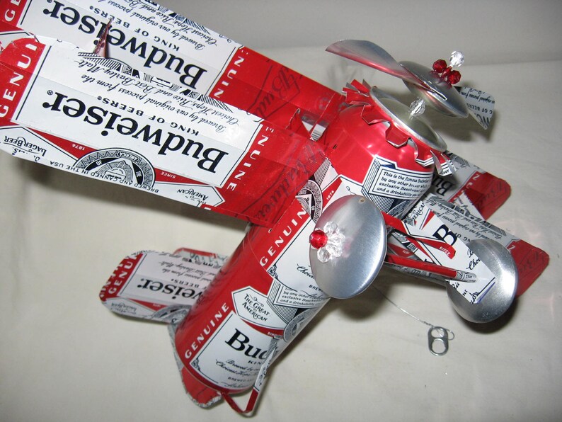 May include: A red, white, and blue Budweiser beer can airplane model with silver wings and propellers. The airplane has red and silver accents and is decorated with rhinestones.