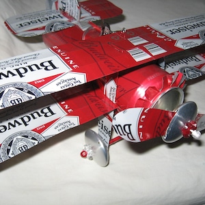May include: A red, white, and blue model airplane made from Budweiser beer cans. The airplane has two sets of wings and a propeller. The wings are made from the top of the cans, and the propeller is made from the bottom of the can.