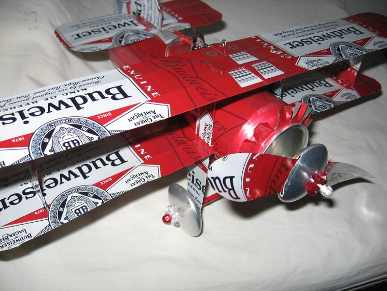 May include: A red, white, and blue model airplane made from Budweiser beer cans. The airplane has two sets of wings and a propeller. The wings are made from the top of the cans, and the propeller is made from the bottom of the can.