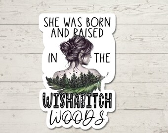 May include: A white sticker with the text "SHE WAS BORN AND RAISED IN THE WISHABITCH WOODS." The sticker features a woman's silhouette with a forest scene on her back and a green feather.