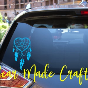 May include: A teal heart-shaped dreamcatcher decal with dangling feathers is displayed on a car window. The words "Bear Made Crafts" are written in yellow cursive below the decal. The car is parked outside.