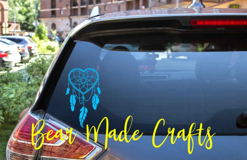 May include: A teal heart-shaped dreamcatcher decal with dangling feathers is displayed on a car window. The words "Bear Made Crafts" are written in yellow cursive below the decal. The car is parked outside.