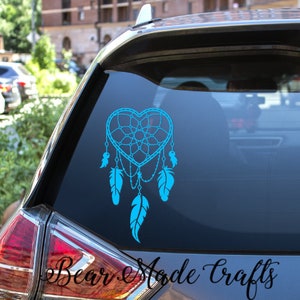 May include: A turquoise heart-shaped dreamcatcher decal with feather accents is displayed on a car window. The decal features a webbed design within the heart shape and dangling feathers. The text "Bear Made Crafts" is at the bottom.