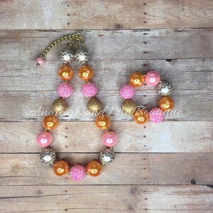 May include: A pink, orange, and gold beaded necklace and bracelet set. The beads are large and have a chevron pattern, glitter, and a shiny finish.