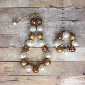 May include: A gold and white chunky bead necklace and bracelet set. The beads are large and have a glittery finish. The necklace has a clasp closure.