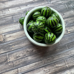 May include: A collection of round, glossy beads featuring a green base with dark green and black striped patterns resembling watermelons. These spherical beads are contained within a smooth, light-colored bowl, showcasing a uniform size and shape.