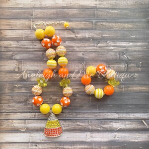 May include: A yellow, orange, and white candy corn necklace and bracelet set. The necklace has a large candy corn charm with rhinestones. The bracelet and necklace are made of large, colorful beads.