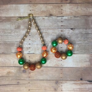 May include: A set of pumpkin-themed necklaces and bracelets. The necklace is made of large, colorful beads in shades of orange, green, and gold. The bracelet is made of smaller beads in the same colors. Both pieces feature sparkly accents.
