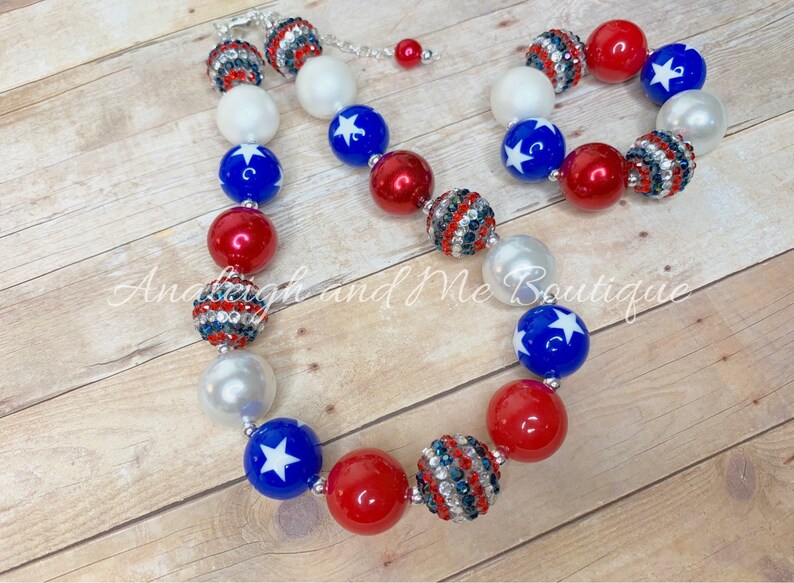 May include: A red, white, and blue beaded necklace and bracelet set with stars and rhinestones. The necklace and bracelet are made of large, chunky beads.