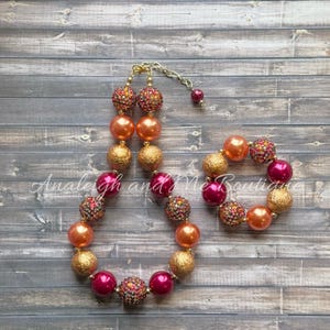 May include: A set of two necklaces with large, colorful beads. The necklaces are made of orange, red, and gold beads with glitter accents. The necklaces are perfect for a little girl.