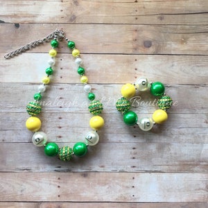 May include: A necklace and bracelet set featuring large, round beads in yellow, green, white, and silver. The necklace has a silver chain and the bracelet is elastic. The beads have a glittery texture and the letter "O" is printed on some of the beads.
