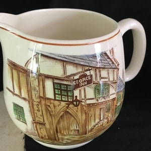May include: A white ceramic creamer with a brown handle and a painted image of a building with the text "The George Inn" on a sign.
