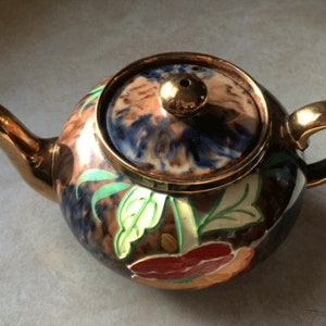 May include: A brown and gold teapot with a floral design. The teapot has a lid with a blue and brown swirl pattern. The teapot is decorated with green leaves and red, orange, and yellow flowers.