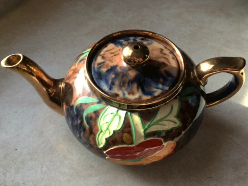May include: A brown and gold teapot with a floral design. The teapot has a lid with a blue and brown swirl pattern. The teapot is decorated with green leaves and red, orange, and yellow flowers.