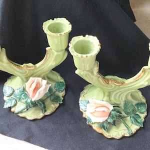 May include: A pair of light green ceramic double candle holders featuring a sculpted pink rose and dark green leaves on the base. Each holder has two cups with scalloped edges and gold-toned trim along the rims and base, showcasing a glossy finish.