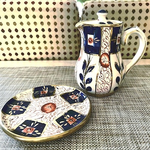 May include: A white and blue ceramic pitcher with a matching saucer. The pitcher has a lid and handle, with a floral and geometric design. The saucer has a similar pattern with a gold rim. The set is on a gray surface.