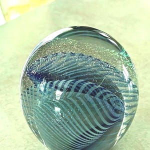 May include: A clear glass paperweight with a blue and white striped design. The sphere has a swirling pattern of blue and white lines, creating a textured, wave-like effect. The glass is transparent, allowing light to pass through.