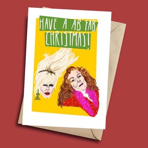 Könnte beinhalten: Weihnachtskarte mit gelbem Hintergrund und dem Text "HAVE A AB FAB CHRISTMAS!". Die Karte zeigt Illustrationen von zwei Frauen mit blonden und braunen Haaren. Eine Frau trägt eine rote Jacke mit einem rosa Pelzkragen.