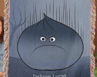May include: A woven throw blanket featuring a dark blue, rounded, grumpy-looking character with wide eyes and a frown. The design includes vertical lines and the name Jackson Lucas printed in white text. The blanket has a fringed edge.