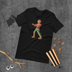 May include: A black short-sleeved t-shirt featuring a graphic of a gingerbread person designed as a zombie. The gingerbread figure wears a torn red top and green pants, with a missing piece on its head and a distressed, ragged appearance.
