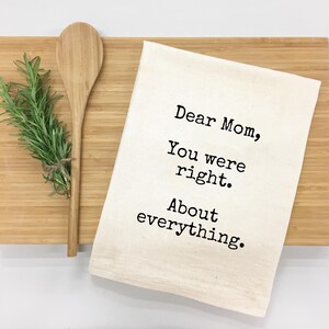 May include: A light-colored kitchen towel with the text "Dear Mom, You were right. About everything." is displayed on a wooden cutting board. A wooden spoon and a bundle of rosemary are also on the board.