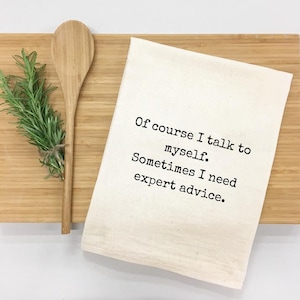 May include: A cream-colored kitchen towel with the text "Of course I talk to myself. Sometimes I need expert advice." is displayed on a wooden cutting board. A wooden spoon and a bundle of rosemary are also present.