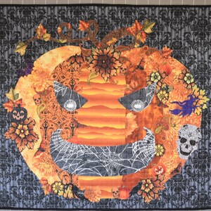 May include: A Halloween-themed quilt featuring a large orange pumpkin with a scary face, surrounded by black spiderwebs, skulls, and fall foliage.