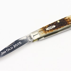 May include: A silver pocket knife with a brown and white handle. The blade is open and engraved with the text "JIM OLD 2015".
