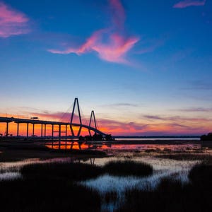 May include: A silhouette of a bridge against a colorful sunset sky. The bridge has a cable-stayed design and is reflected in the water below. The sky is a mix of pink, orange, and blue.