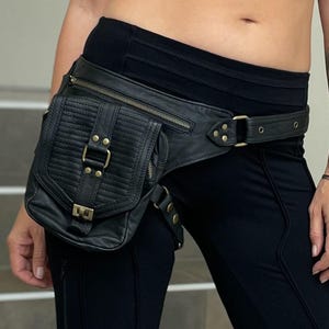 May include: Black leather fanny pack with a rectangular, quilted front pocket and a zippered main compartment. The pack features brass-colored hardware and a belt-style strap. The wearer is also wearing black leggings and a black crop top.