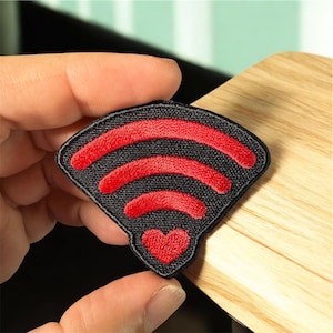 May include: A black and red embroidered patch featuring a Wi-Fi symbol with a heart at the bottom. The patch has a black background with three red curved lines and a red heart. The patch is held by a person's fingers.