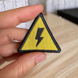 May include: Yellow triangle warning sign with a black lightning bolt inside. The sign is embroidered with black thread.