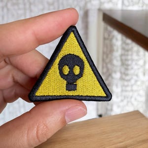 May include: Yellow triangle warning sign with a black skull and crossbones graphic. The sign is embroidered with black thread.